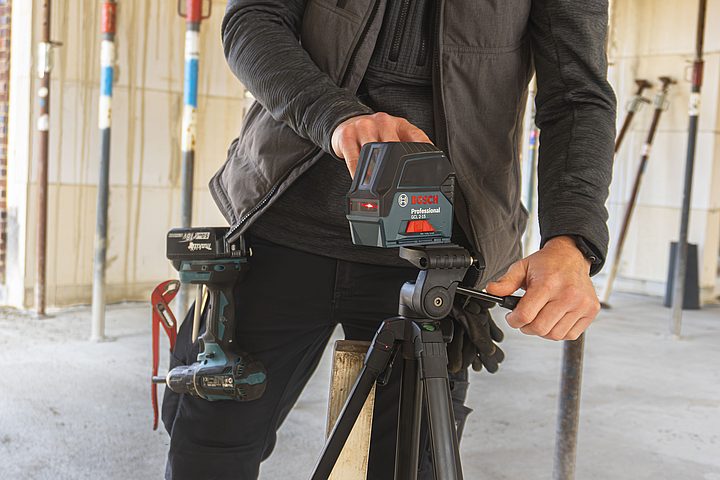 Ein Mitarbeiter auf einer Baustelle bedient ein motorisiertes Arbeitsgerät auf einem Stativ. Er trägt Arbeitskleidung und Werkzeug am Gürtel; im Hintergrund ist ein Rohbau zu sehen.