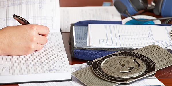 Eine Person schreibt in ein aufgeschlagenes Formularbuch, daneben liegen Schreibutensilien und ein Navigationsrechner.